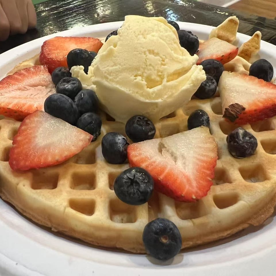 Belgian waffle topped with vanilla ice cream, sliced strawberries, blueberries, and wafer pieces on a white plate.