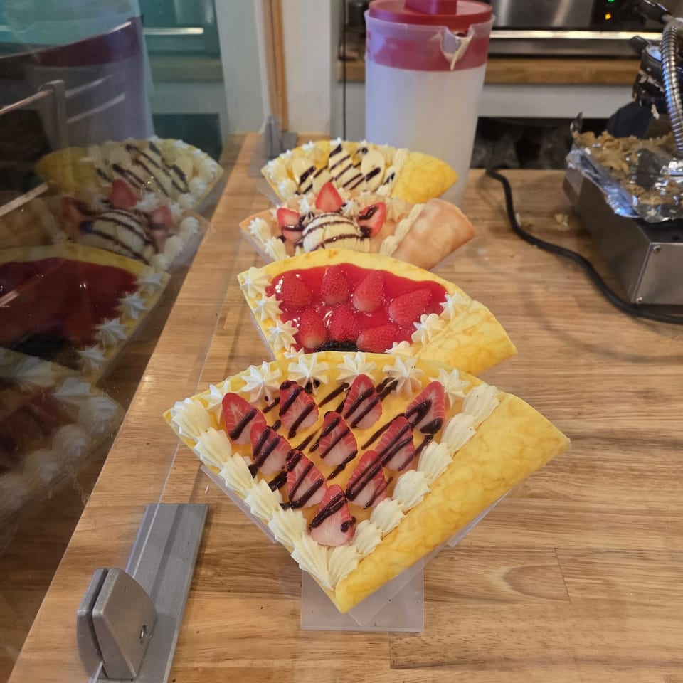 Assorted fruit-topped dessert crepes with whipped cream and chocolate drizzle displayed on a wooden restaurant counter.