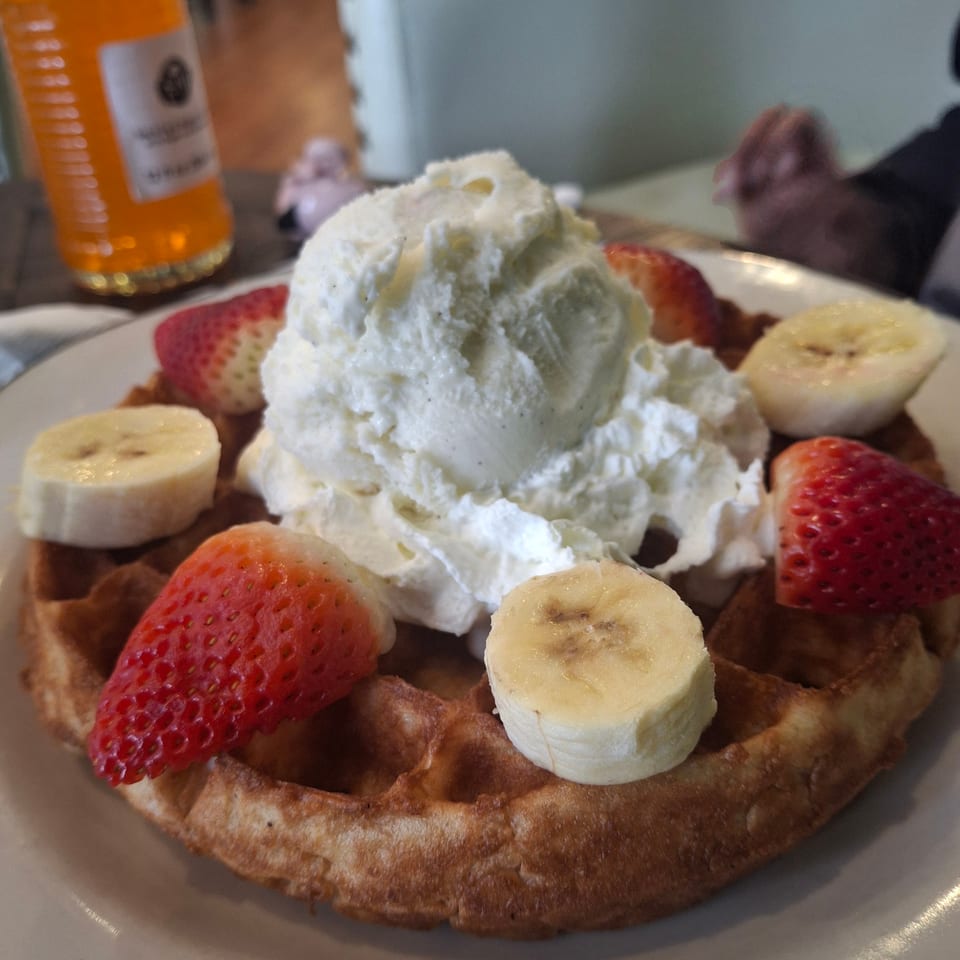 Golden waffle topped with whipped cream, sliced strawberries and bananas, served on a plate beside an orange drink.