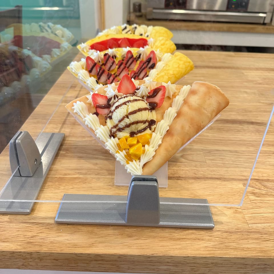 Dessert crepes topped with whipped cream, strawberries, mango, ice cream, and chocolate sauce displayed on a café counter.