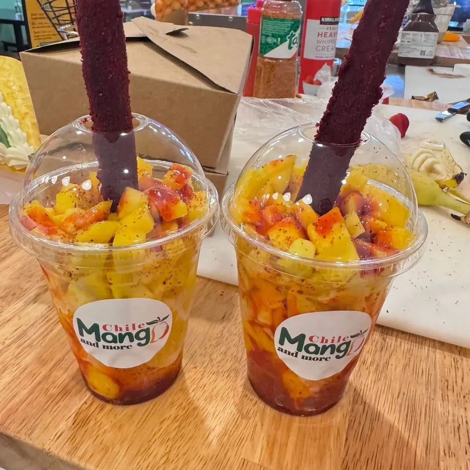 Two cups of chopped mango and fruit topped with chili seasoning and tamarind sticks on a restaurant counter.