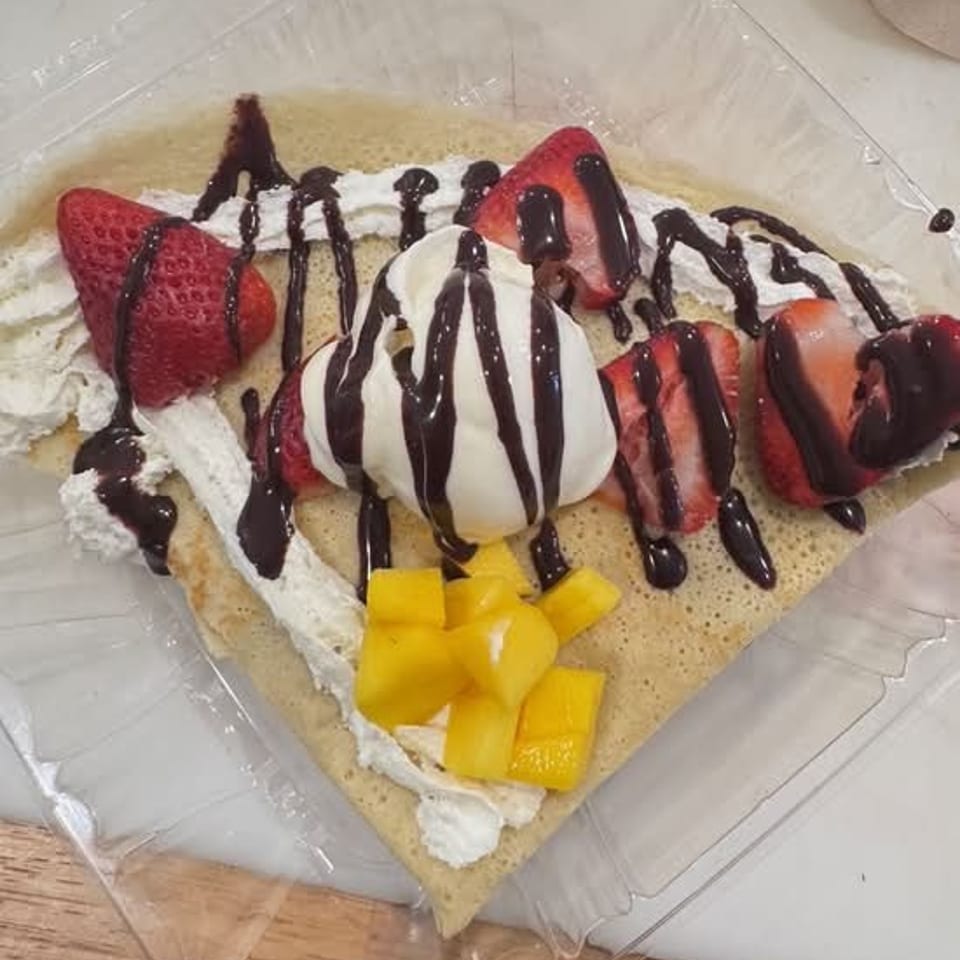 Crepe topped with vanilla ice cream, strawberries, mango chunks, whipped cream, and drizzled chocolate sauce in a takeaway container.